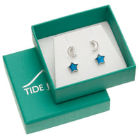 A pair of silver-toned stud earrings displayed in an open teal gift box. One earring is a crescent moon and the other is a five-point star with bright blue enamel inlay.